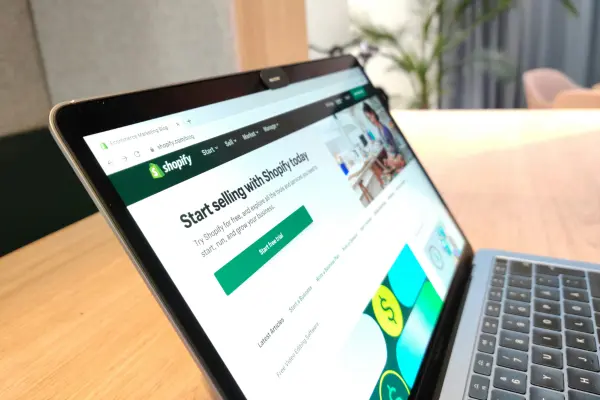 A laptop displaying the Shopify start page on its screen, placed on a wooden table in a modern workspace. The screen headline reads 'Start selling with Shopify today', illustrating the process of setting up an online store, suitable for an informative guide about IOSS registration and customs management.