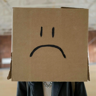 Person wearing a cardboard box with a large sad face drawn on it, symbolizing disappointment or dissatisfaction with a mystery box purchase.
